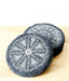 Two round black slate coasters with intricate white mandala patterns, stacked on a light wood surface against a white background.