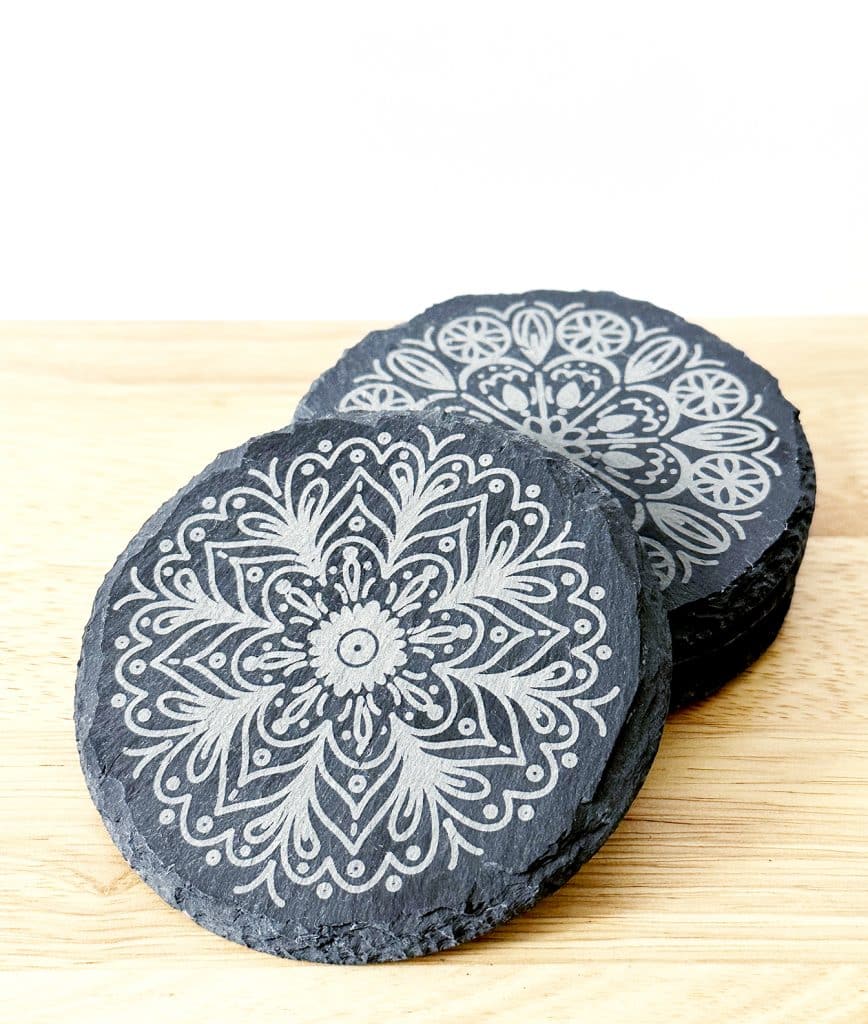 Two round black slate coasters with intricate white mandala patterns, stacked on a light wood surface against a white background.