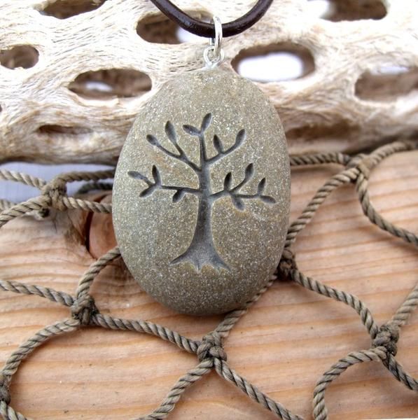 A tree-of-life laser engraved stone pendant on a dark cord necklace, displayed on a rustic wooden surface with rope and driftwood.