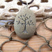 A tree-of-life laser engraved stone pendant on a dark cord necklace, displayed on a rustic wooden surface with rope and driftwood.